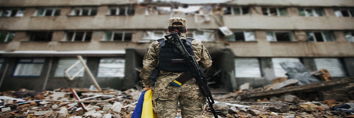 Ukrainische Militärfrau mit ukrainischer Flagge in ihren Händen vor dem Hintergrund eines explodierten Hauses					