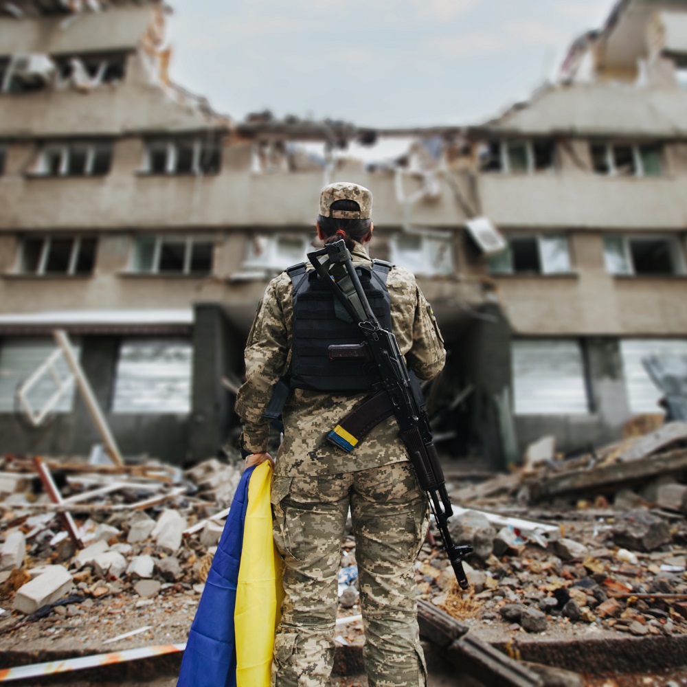 Ukrainische Militärfrau mit ukrainischer Flagge in ihren Händen vor dem Hintergrund eines explodierten Hauses					