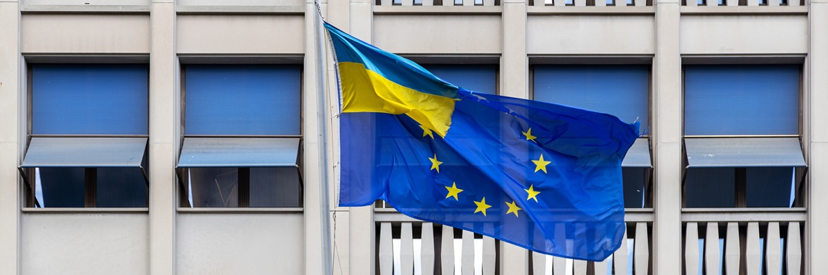 Ukraine and EU flags waving together outside building. A combined Ukraine and European Union flag waves in front of a modern office building, symbolizing political unity and support.